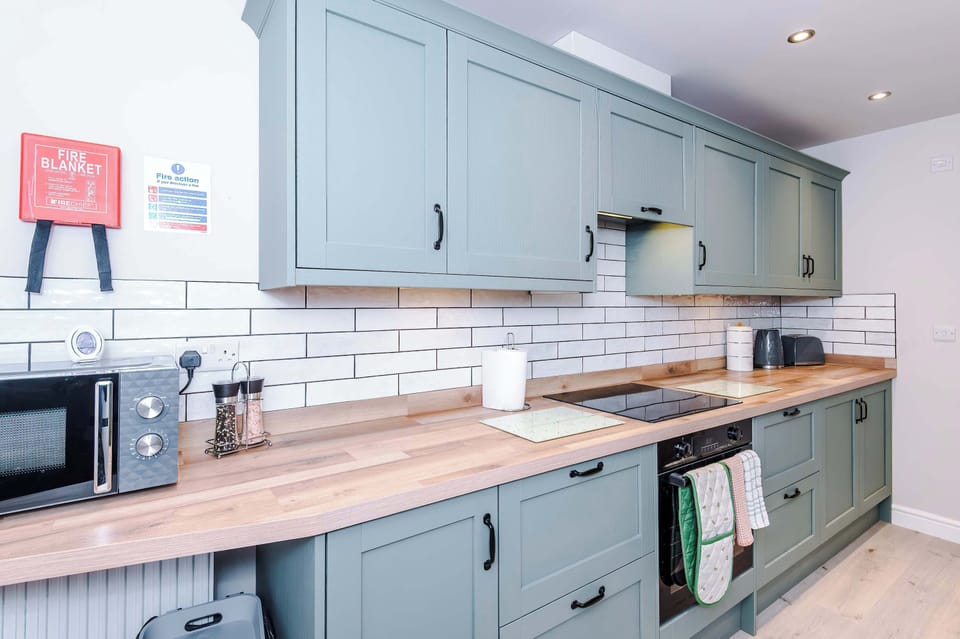 A sleek modern kitchen with light gray cabinets, wooden countertops, white subway tile backsplash, and neatly arranged appliances including a microwave, toaster, kettle, and knife block, creating a clean and functional cooking space.