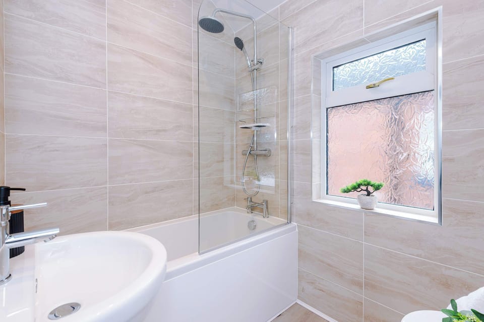 A sleek modern bathroom with beige marble tiles, a white bathtub and glass shower screen, chrome fixtures, and a bright frosted window with a potted plant, creating a clean and stylish space.