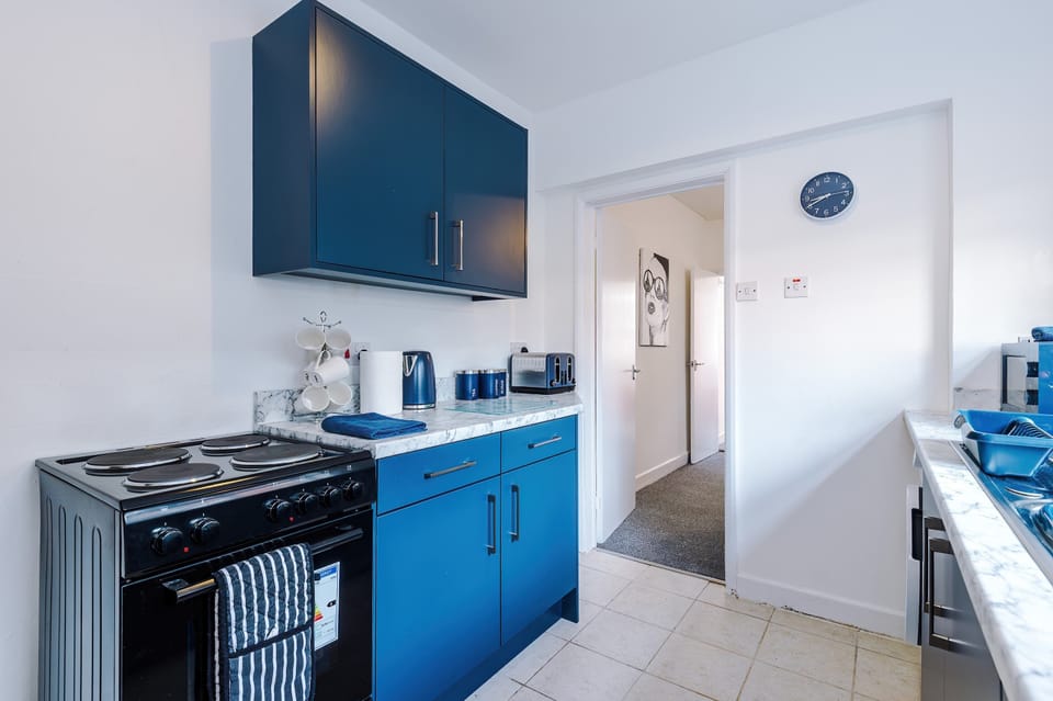 A sleek kitchen with blue and white tones, marble counters, and tiled floors. Built-in washing machine, coffee machine, and fruit bowl add functionality, while natural light creates a fresh, inviting atmosphere.