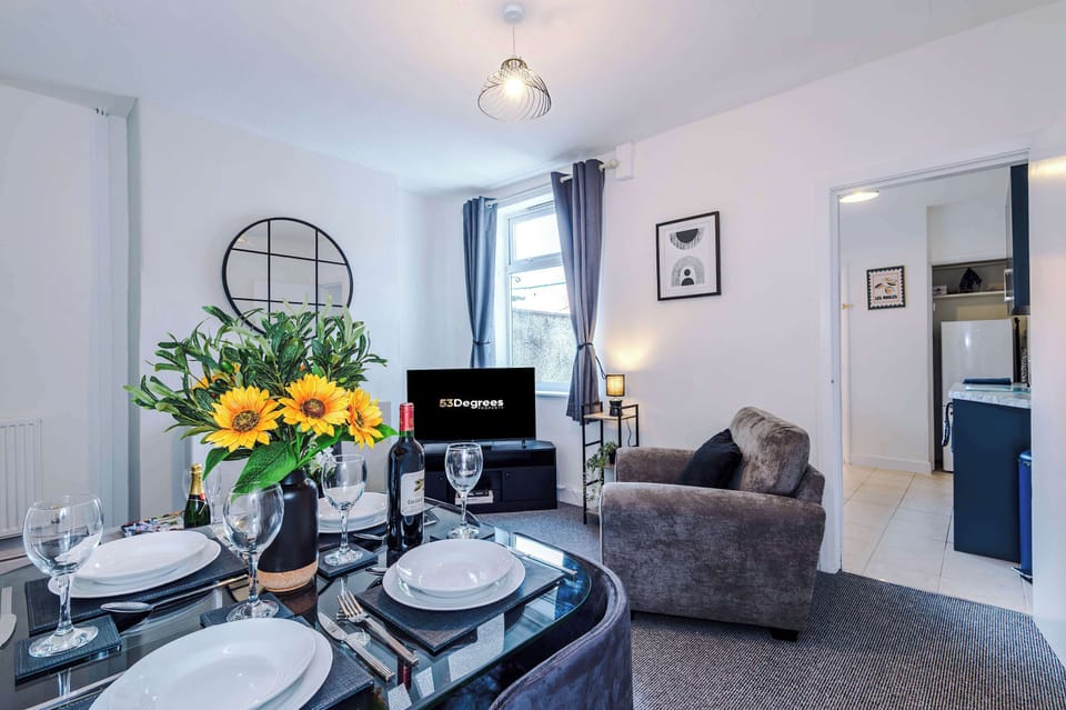 - Modern living at its finest: gray sofa and armchair, fireplace with round mirror, glass coffee table, and a dining table set with wine and sunflowers, all framed by natural light and greenery.
