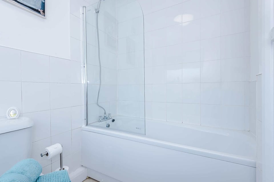 A clean, minimalist bathroom with tiled walls, pedestal sink, turquoise towels, and framed art. A window with a light blue blind and warm lighting enhance the fresh, welcoming vibe.