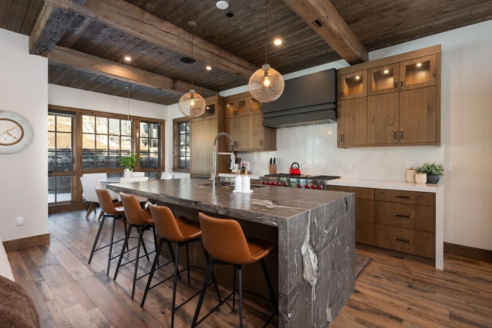 Open kitchen with large stone waterfall island, bar seating, wood cabinetry, and reclaimed beam ceiling