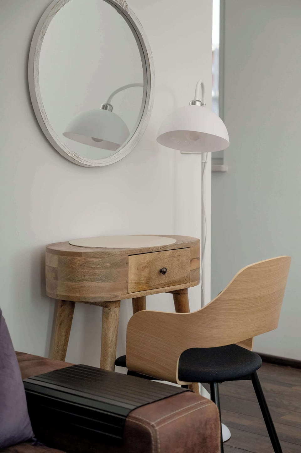 A vertical photo of a stylish, modern vanity table with a round mirror, a wooden chair, and an pendant light in a clean, white room.