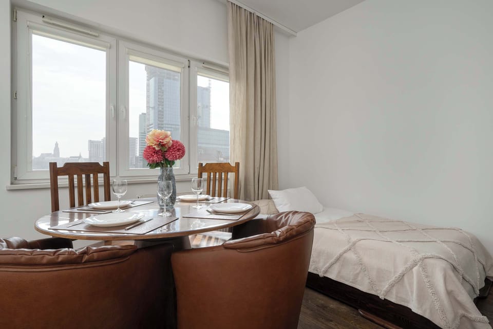 A view across the living area, showing a round dining table and four chairs in front of a window, adjacent to a single bed with white linens.