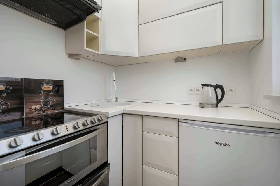 A compact kitchen corner featuring a small range, white lower cabinets, a small refrigerator, a kettle, and white upper cabinets.
