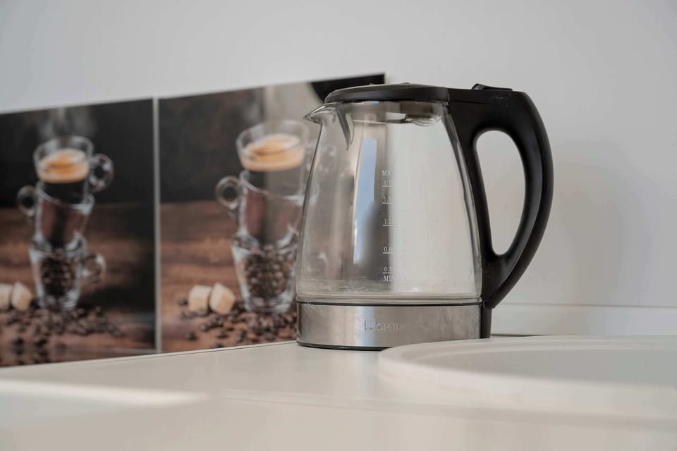 A close-up of kitchen items on a white countertop, including a glass electric kettle and a small, square framed picture of coffee cups.