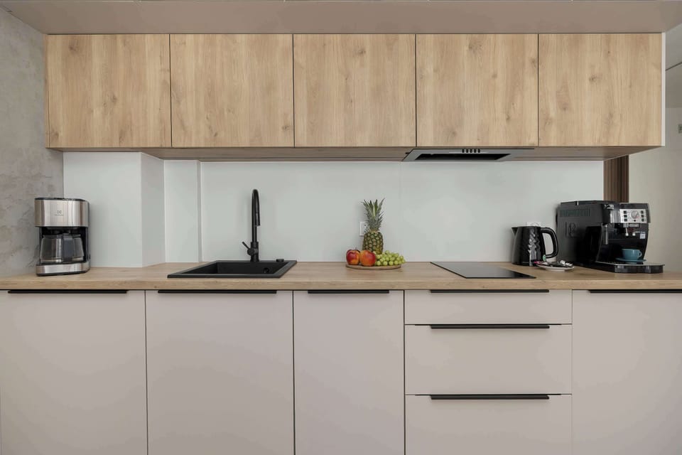 A modern kitchenette with light beige cabinetry, wooden upper units, and built-in appliances including a coffee machine and kettle.