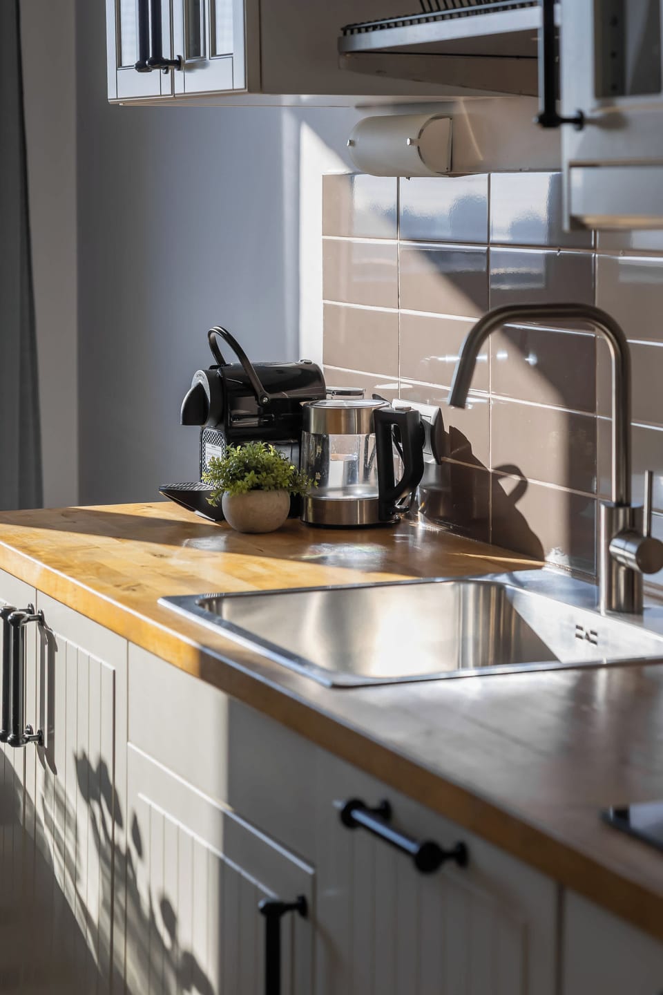 A kitchen countertop detail highlights a stainless steel sink and a modern black faucet. A small black espresso machine and electric kettle are conveniently placed on the wooden surface.