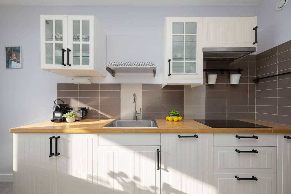 A classic white kitchen is equipped with handleless cabinetry, a light wood countertop, and a tiled backsplash. The compact design includes a built-in oven and a modern sink area.