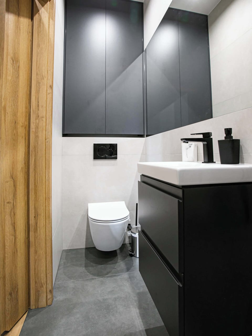 A bathroom corner featuring a toilet and vanity with a mirror above it. The design remains consistent with gray tiles, wooden elements, and modern fittings, emphasizing practicality and style.