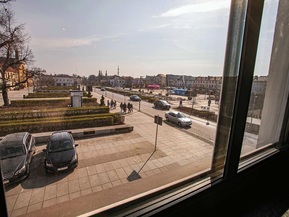 A view from the window overlooking a street with parked cars and nearby buildings. The perspective captures a calm neighborhood scene with a mix of residential and urban surroundings.