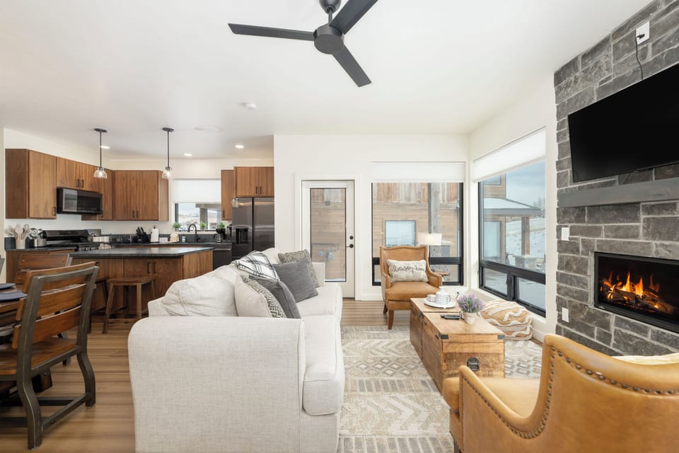 Relax in your open-concept haven at Base Camp Townhome 106 where modern kitchen flows into cozy living space, complete with crackling fireplace and warm wood accents.