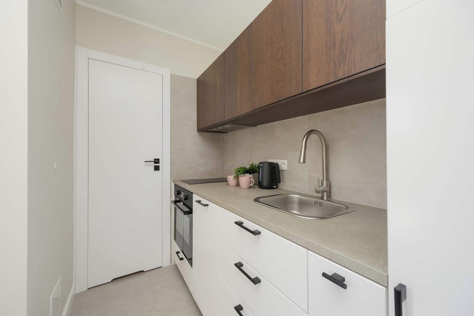 A wide-angle view of the kitchenette displays the contrast between the dark overhead cabinetry and the white base drawers. A white door on the left leads further into the apartment.
