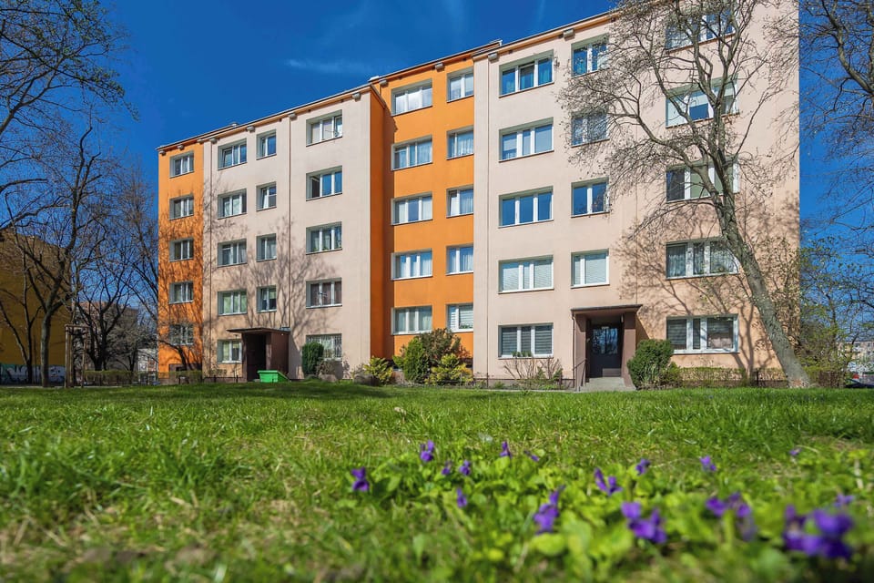 The exterior of the apartment building is shown as a multi-story block with a facade of white and orange panels. A lush green lawn and small purple flowers in the foreground highlight the building's surrounding greenery.