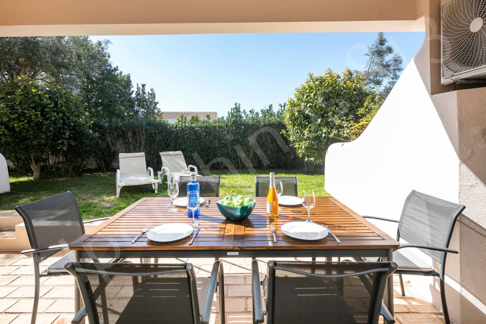 Al fresco dining setup on the covered terrace at 813 Alporchinhos with a wooden table laid for dinner with plates, wine glasses, bottles of wine and water, surrounded by the private garden with sun loungers on a lush green lawn, perfect for evening meals in the Algarve sunshine