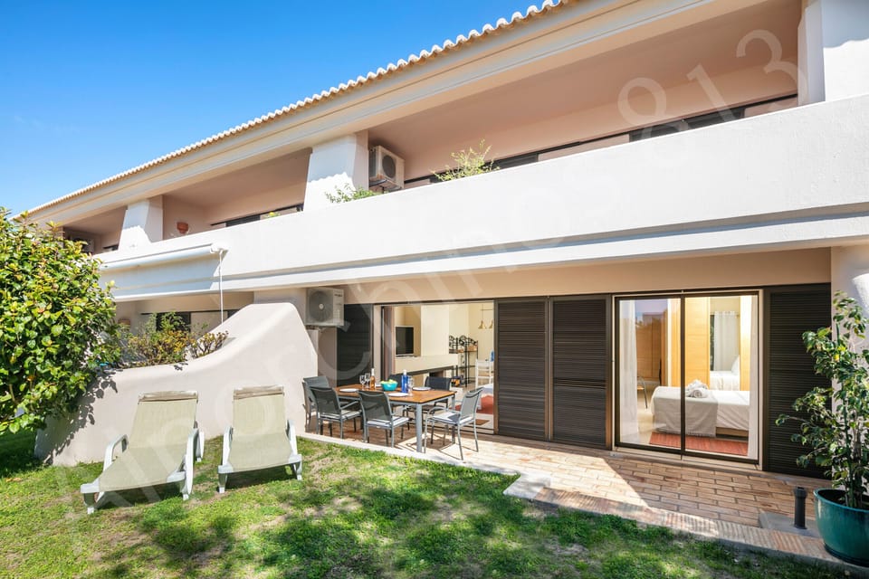 Private garden terrace at 813 Alporchinhos holiday home featuring an outdoor dining table with chairs under a covered pergola, white sun loungers on a manicured lawn, and lush green landscaping surrounding the ground-floor apartment in this popular Algarve resort