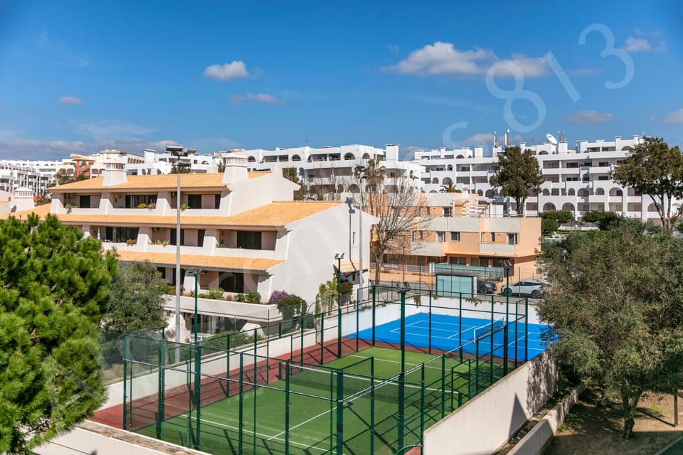 Elevated view of the Alporchinhos resort complex near 813 Alporchinhos showing the white and terracotta apartment buildings, a tennis court, communal swimming pool, and beautifully landscaped gardens with mature trees, all set beneath clear blue Algarve skies