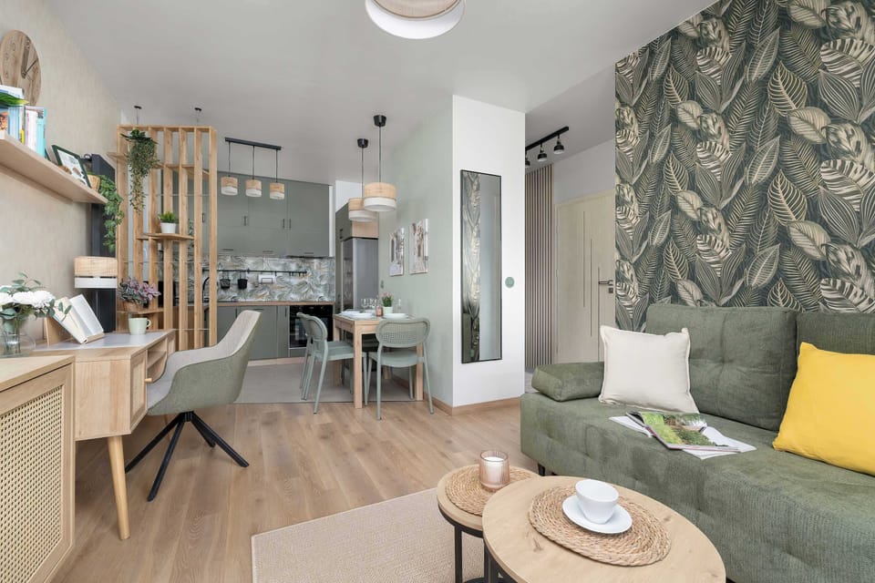 A bright open-plan space combining a dining area and living room, with a wooden table and upholstered chairs. Large windows and patterned curtains bring in natural light, enhancing the airy feel of the interior.