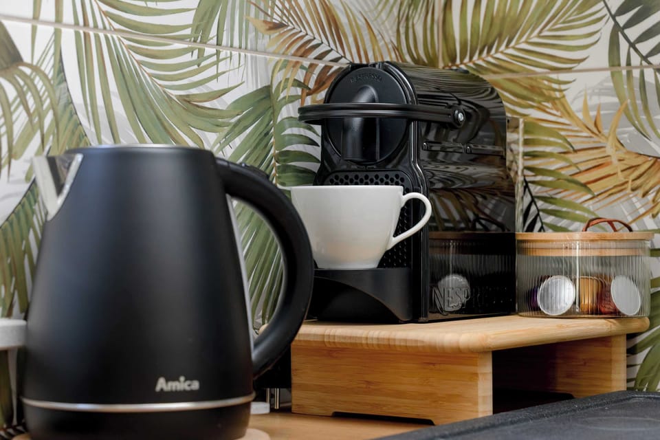 A coffee station set on a wooden countertop with a capsule coffee machine, an electric kettle, and cups neatly arranged. A tropical-patterned backsplash adds a vibrant and stylish touch to the setup.
