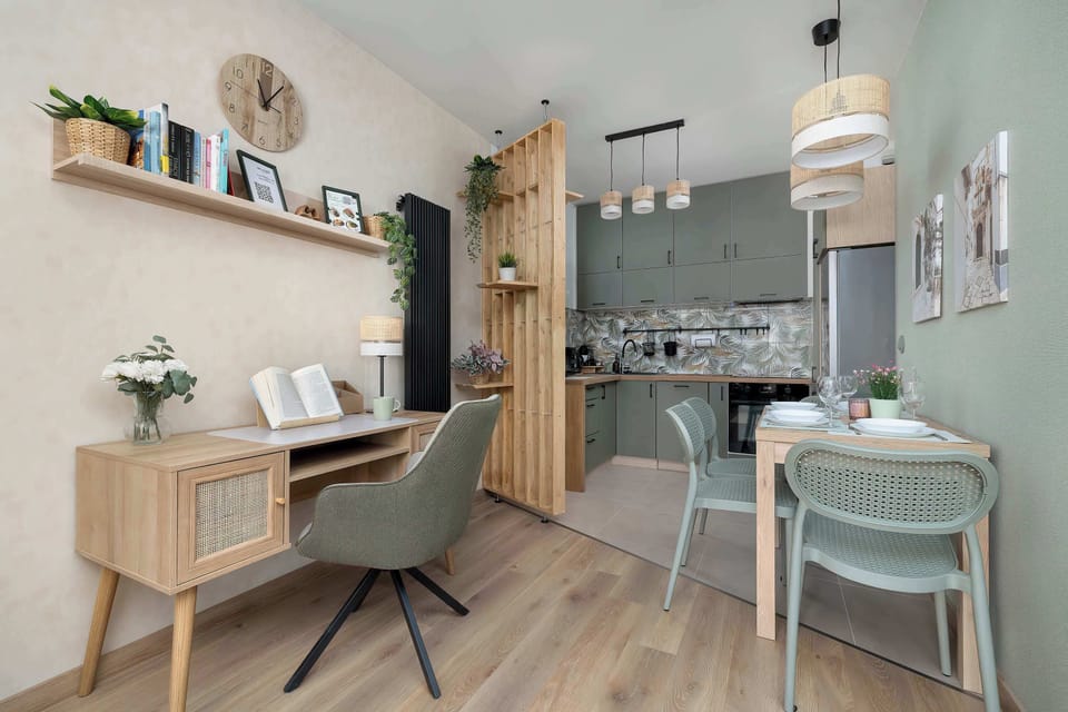 A bright dining area with a wooden table and upholstered chairs placed next to a small workspace with a desk and chair. Light wood finishes and soft green accents create a cohesive, modern look, while decorative shelves add a homely touch.