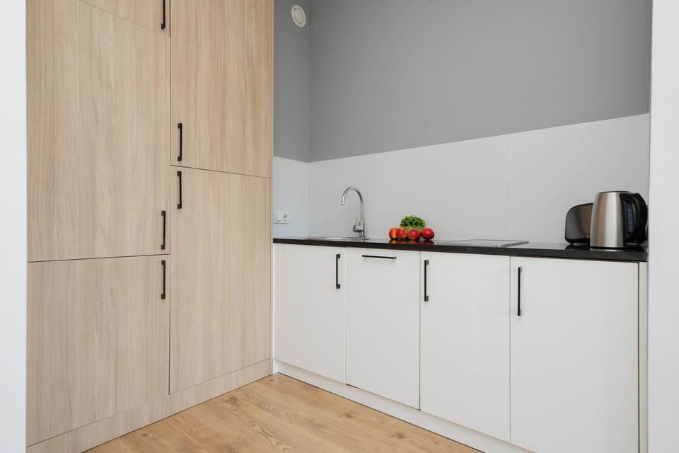 A modern, light-filled kitchenette with simple white lower cabinets and matching tall wooden storage cabinets. The floor is light wood.