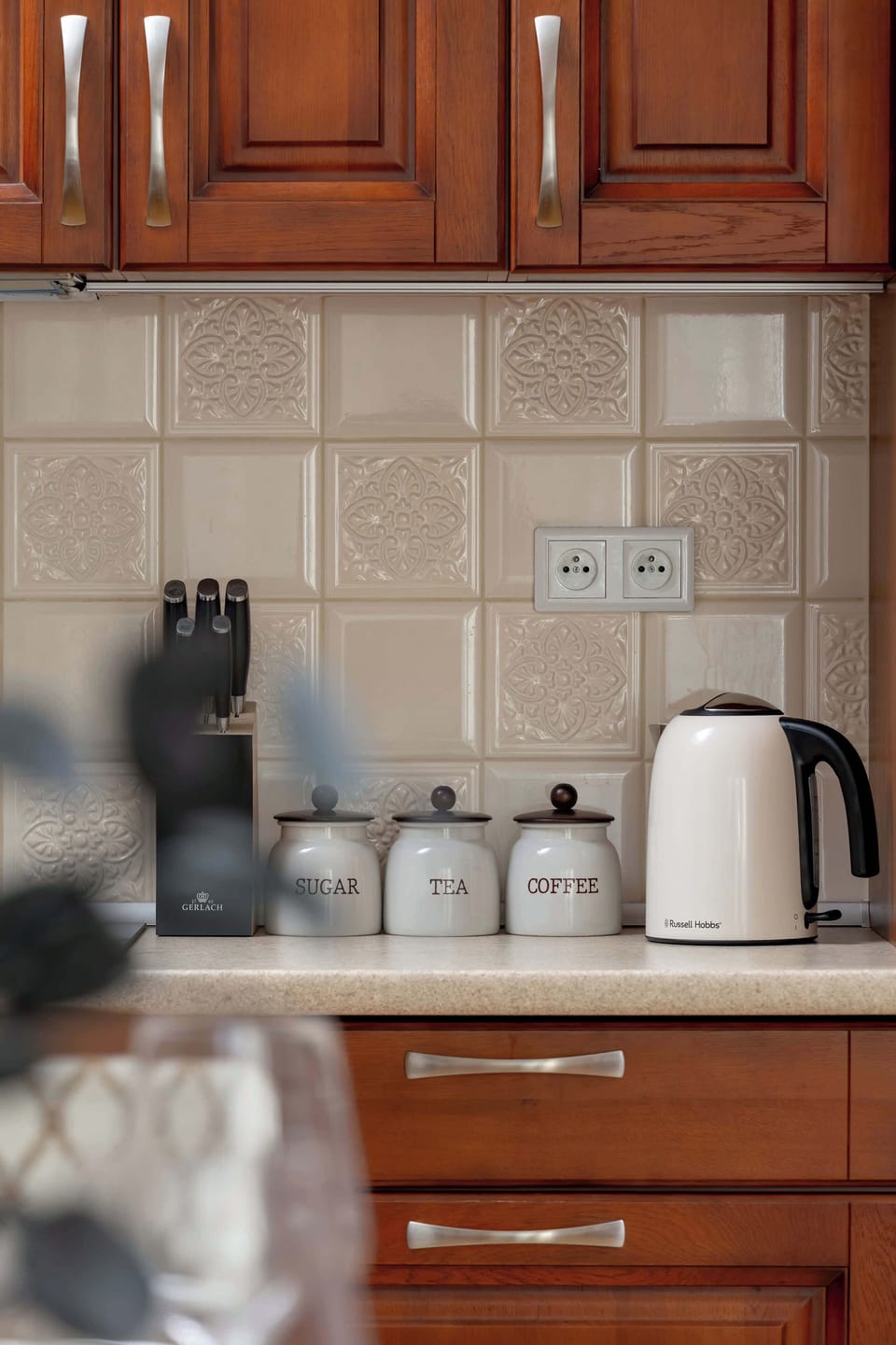 A close-up shot of the kitchen counter shows a white electric kettle and essential cooking utensils neatly organized. The tiled backsplash is easy to clean and matches the neutral tones of the room.