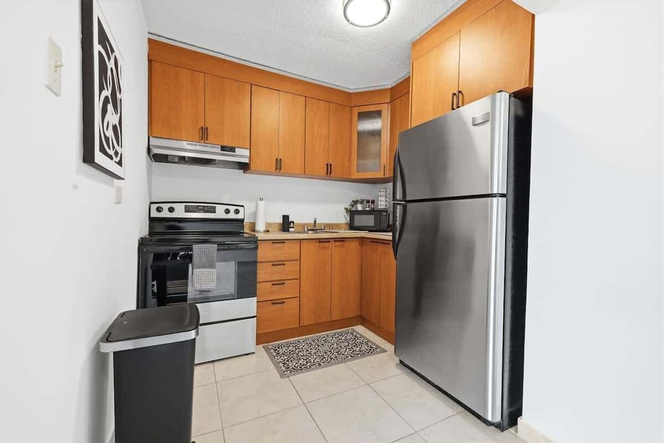 Functional kitchen with L-shaped wood cabinets, stainless-steel refrigerator and stove, light countertops, and a clean tiled floor, creating a bright and practical cooking space.