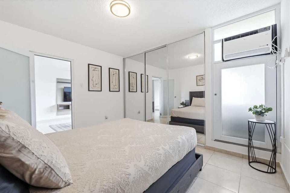 Light-filled bedroom with a queen bed, mirrored closet doors that reflect the space, minimalist wall art, and a window with natural light and an AC unit above.