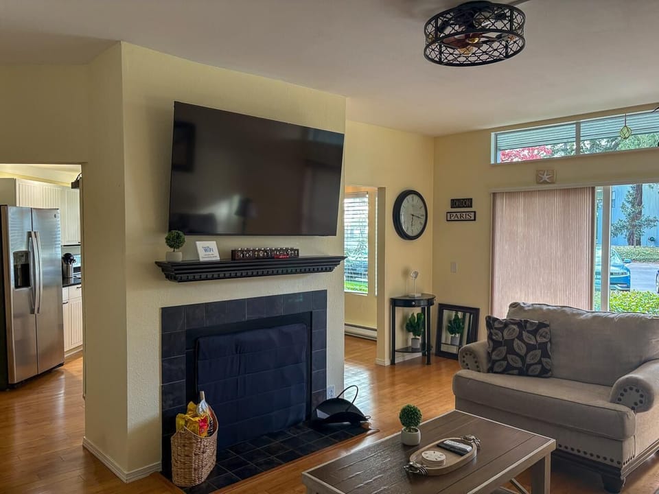 Flat-screen TV mounted above a black-tiled fireplace, with a gray loveseat and wood coffee table nearby.