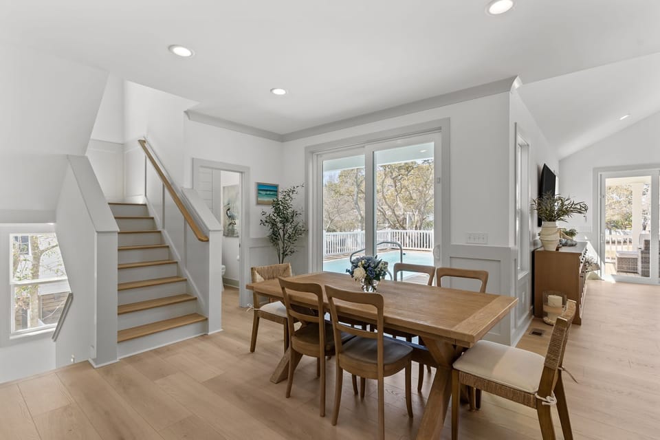 Open dining space with a wood table, six chairs, blue floral centerpiece, light wood floors, and a bright white kitchen with island seating, pendant lights, stainless appliances, and nearby hallway doors.