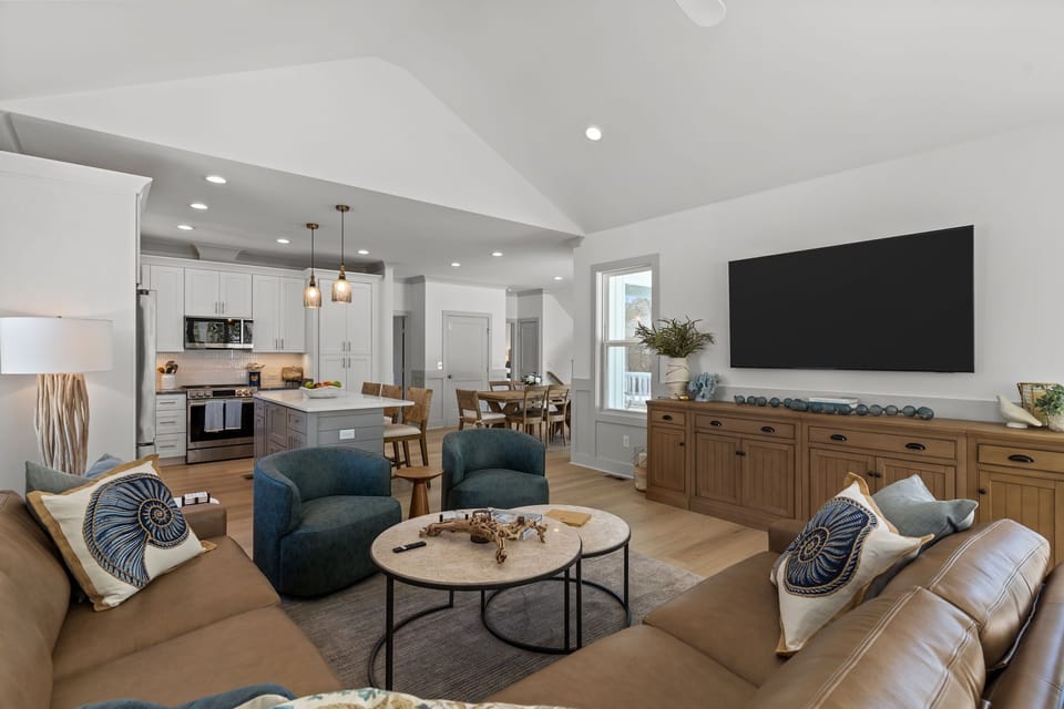 Bright living room with vaulted ceiling, leather sectional, two blue chairs, round coffee tables, wood console, large wall TV, coastal decor, and sliding glass doors opening to a covered deck.