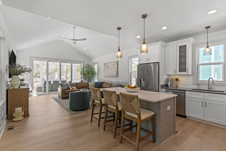 Bright white kitchen with quartz island, three woven barstools, pendant lights, stainless fridge, microwave, stove, sink, tall cabinets, wood floors, and a fruit bowl in this stylish beach house kitchen.