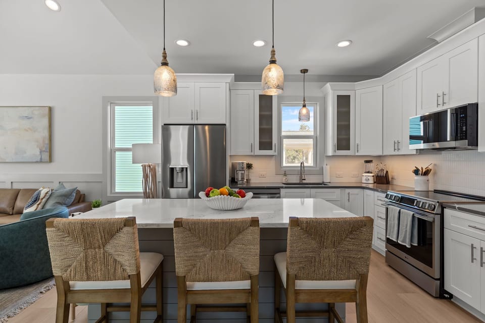 Bright white kitchen with quartz island, three woven barstools, pendant lights, stainless fridge, microwave, stove, sink, tall cabinets, wood floors, and a fruit bowl in this stylish beach house kitchen.