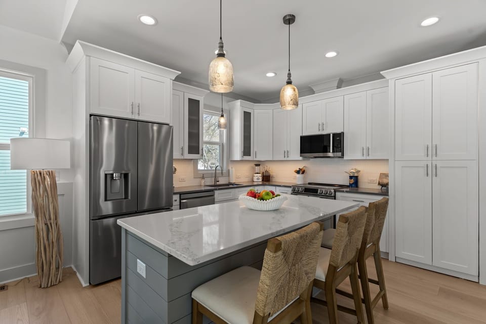 Bright white kitchen with quartz island, three woven barstools, pendant lights, stainless fridge, microwave, stove, sink, tall cabinets, wood floors, and a fruit bowl in this stylish beach house kitchen.