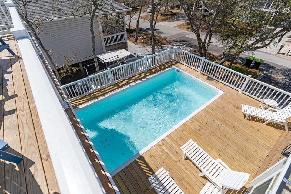 Overlook of the private pool, wood sun deck, white lounge chairs, and white railing from the upper balcony, with quiet neighborhood trees and homes surrounding the sunny backyard.