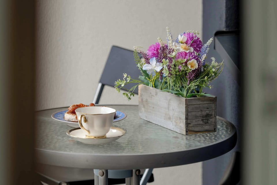 A small round bistro table on the balcony holds a cup of coffee and a wooden box filled with flowers. The blurred background suggests a peaceful outdoor setting.