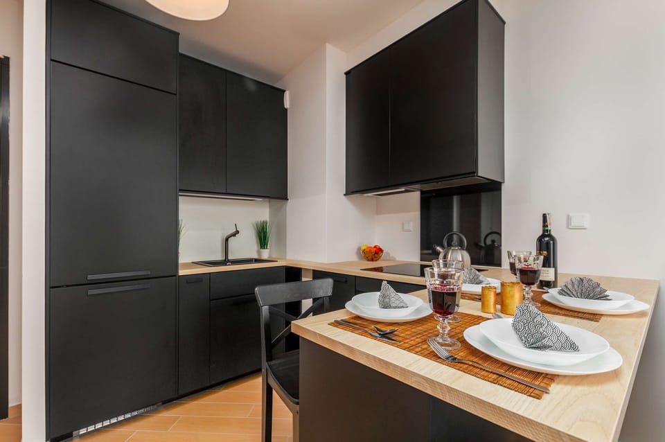 This sleek kitchenette area showcases black minimalist cabinetry and a wooden dining counter. The counter is set with plates, napkins, and wine glasses for a meal.