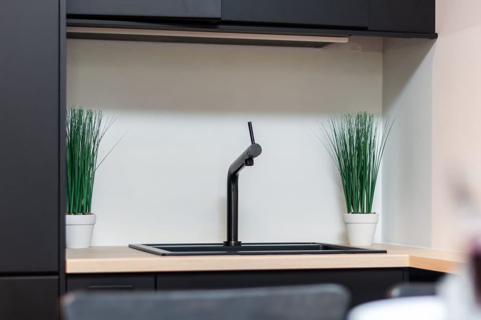 A minimalist black kitchen faucet is paired with a dark sink on a light wood countertop. Two small pots of grass sit on either side, adding a fresh element to the workspace.