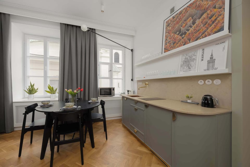 A bright, modern kitchen and dining area features sleek grey cabinetry, black dining chairs, and a round black table. Hardwood parquet flooring and a subtle wall print complete the space.