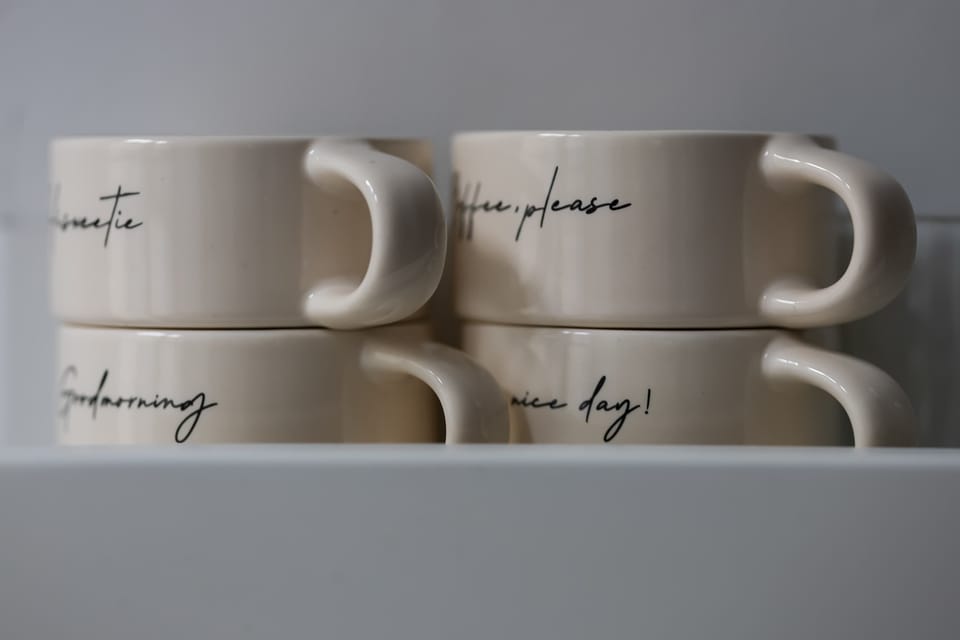 Four elegant white ceramic mugs with delicate gold rims and decorative scripts are neatly stacked in two rows, resting on a flat surface.