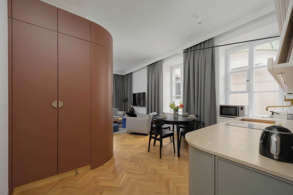 A modern kitchen and dining space with minimalist cabinetry and a dark dining table is bordered by a distinct curved, terracotta-colored wall structure. Parquet flooring and pendant lights define the ambiance.