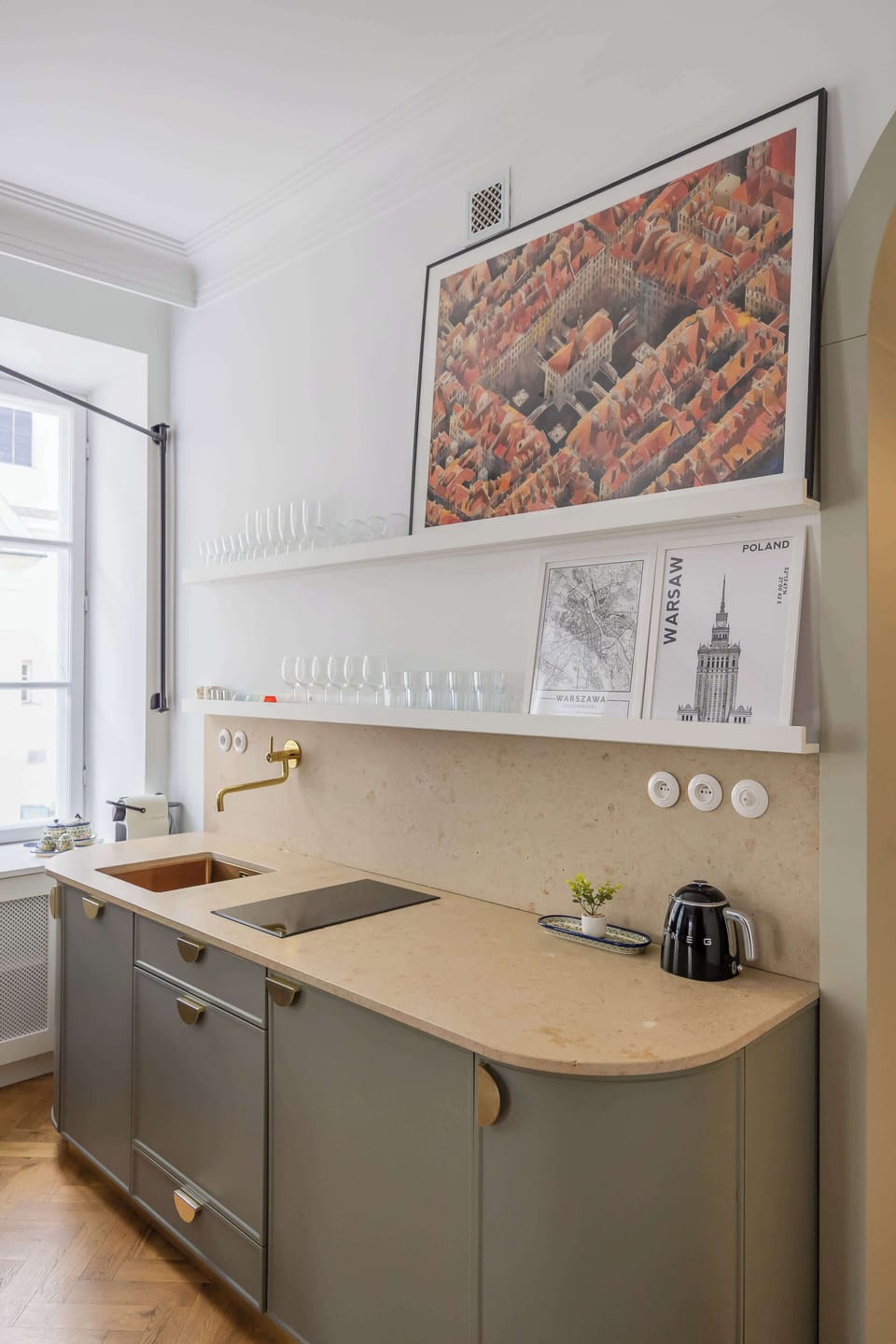 A view into a contemporary kitchen shows minimalist light grey cabinetry, a compact countertop with a small induction cooktop, and a patterned wall art panel near a window.