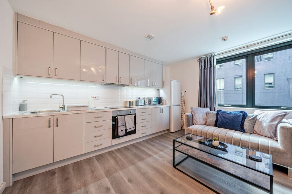 Open-plan kitchen with fitted cupboards, sink, oven, fridge-freezer, and a sofa beside a large window with curtains.