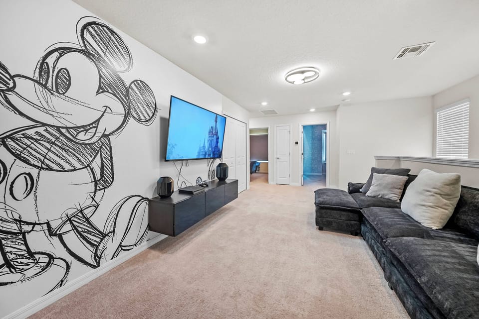 Discover a playful lounge with a vibrant Mickey Mouse mural that adds whimsy. Relax on the plush dark sectional, enjoy the cozy carpet, and soak in the modern lighting—an ideal spot to unwind after your adventures.