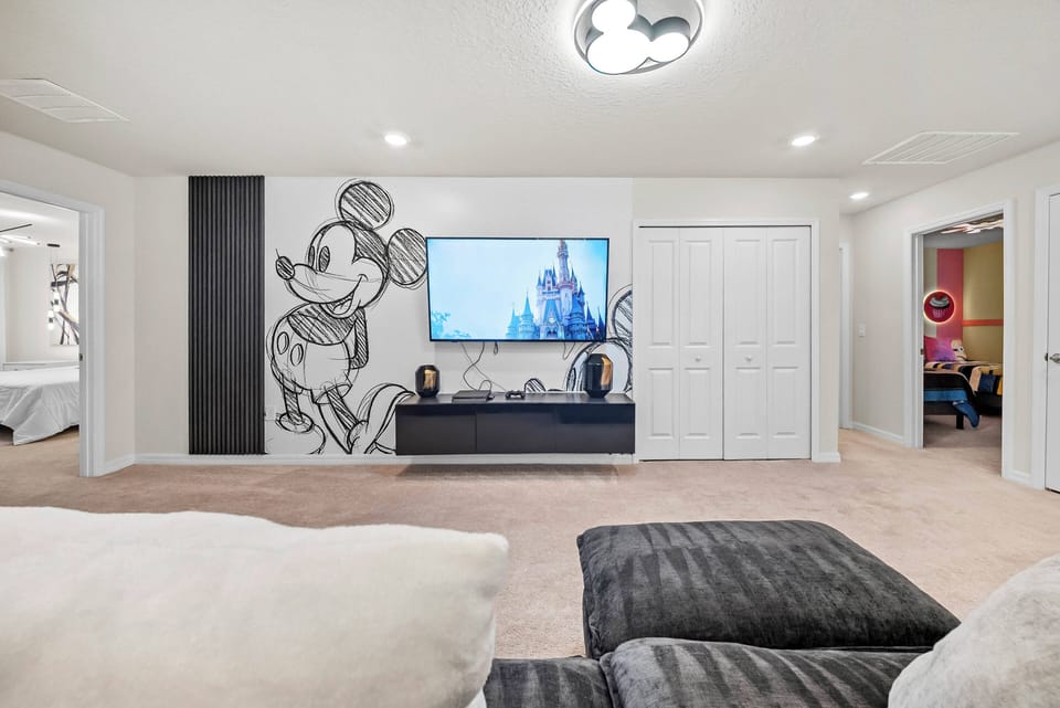 Step into a playful living area adorned with a whimsical Mickey Mouse mural, perfect for family fun. The plush carpet and cozy sectional invite you to unwind, while the large television sets the stage for movie nights together.