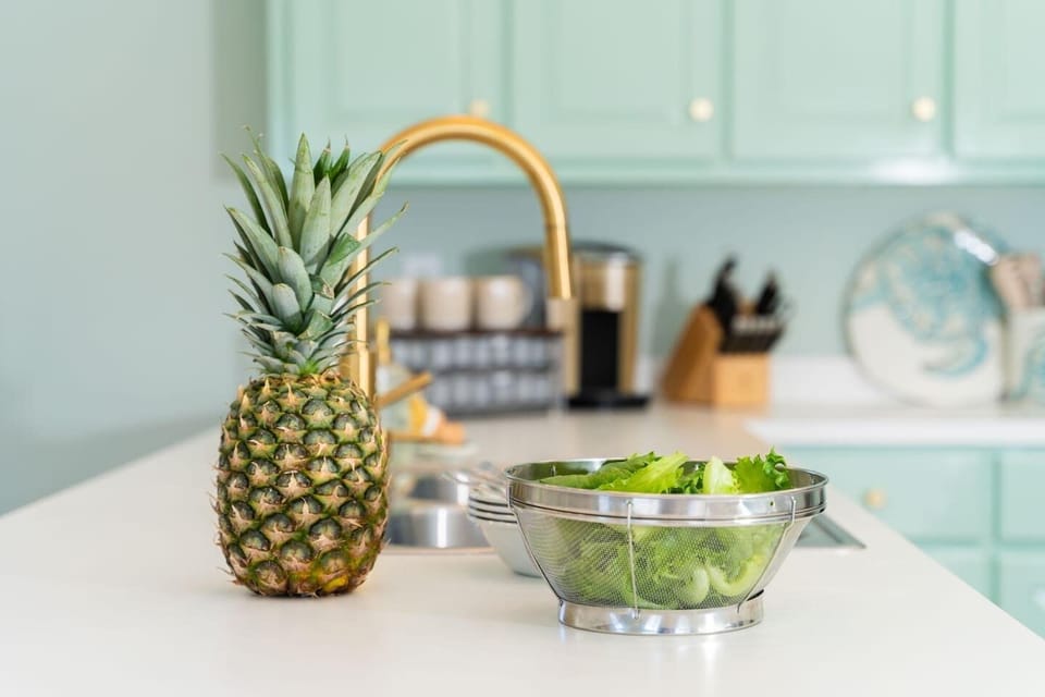 A close-up of the kitchen showcases thoughtful styling with a fresh pineapple and crisp greens in the foreground, highlighting the home’s clean, inviting aesthetic. Paired with functional prep elements, this space blends coastal charm with everyday u