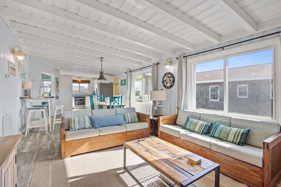 Bright coastal living room with sofas, chairs, TV, coffee table, floor lamp, wood ceiling beams, and large windows plus a door opening to a deck with ocean views.
