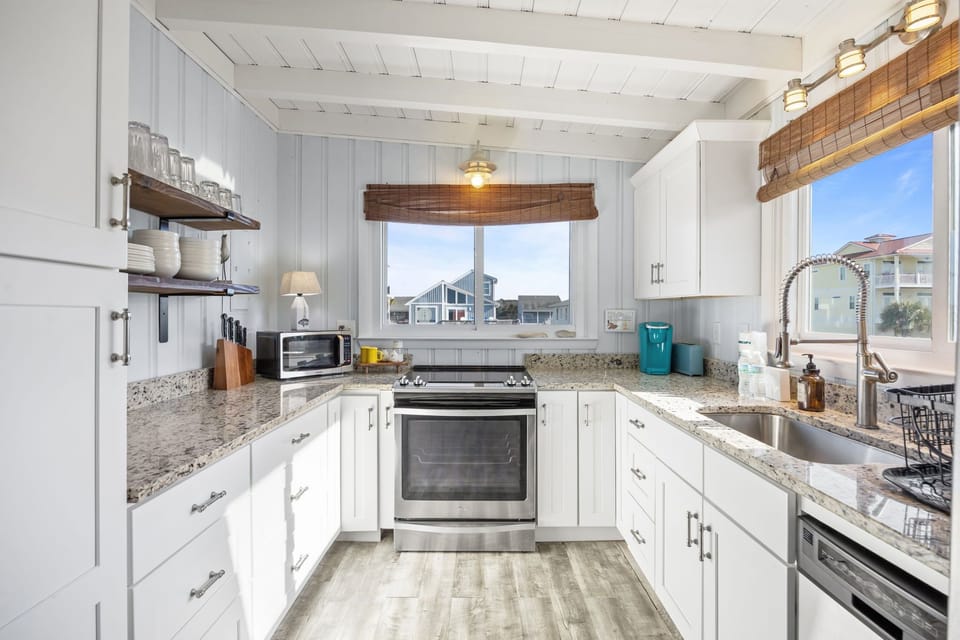 Bright white kitchen with granite counters, stainless stove and dishwasher, deep sink, open shelves, wood-look floors, and sunny windows with neighborhood views.