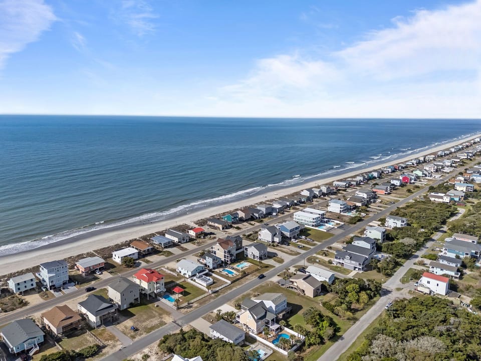Aerial view of an oceanfront Oak Island beach house marked near the shoreline, with nearby homes, quiet streets, sandy beach, and wide Atlantic Ocean views.
