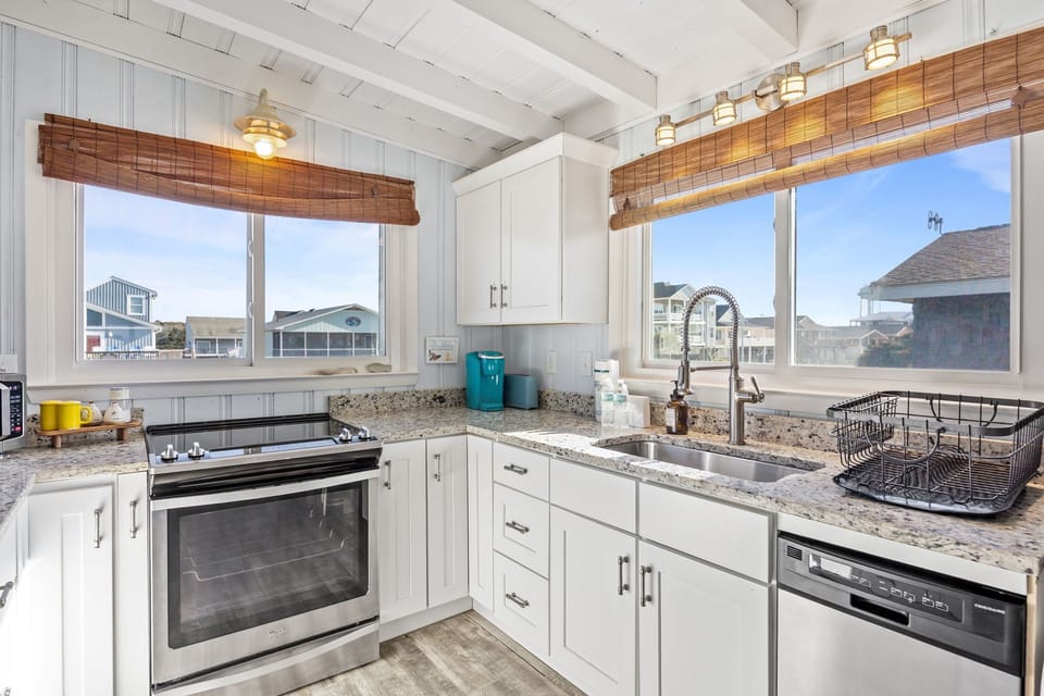 Bright white kitchen with granite counters, stainless stove and dishwasher, deep sink, open shelves, wood-look floors, and sunny windows with neighborhood views.